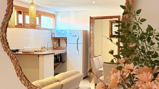 A round mirror with a woven frame on a wall, reflecting a modern kitchen with a refrigerator, a sofa, and a dining area. - Apartamento Messina  (Jijoca de Jericoacoara)