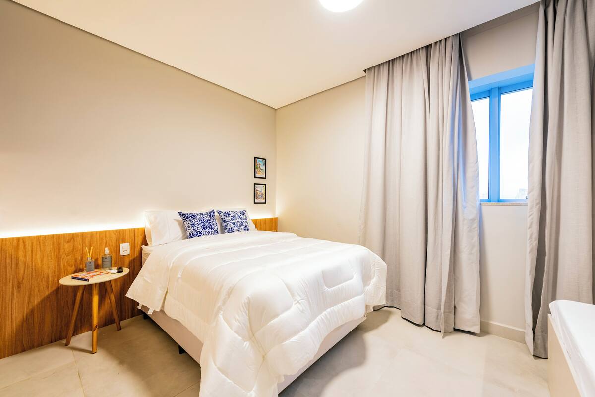 A hotel room with a large bed, a bedside table with a lamp, a small table with a book and a bottle, and a window with curtains.