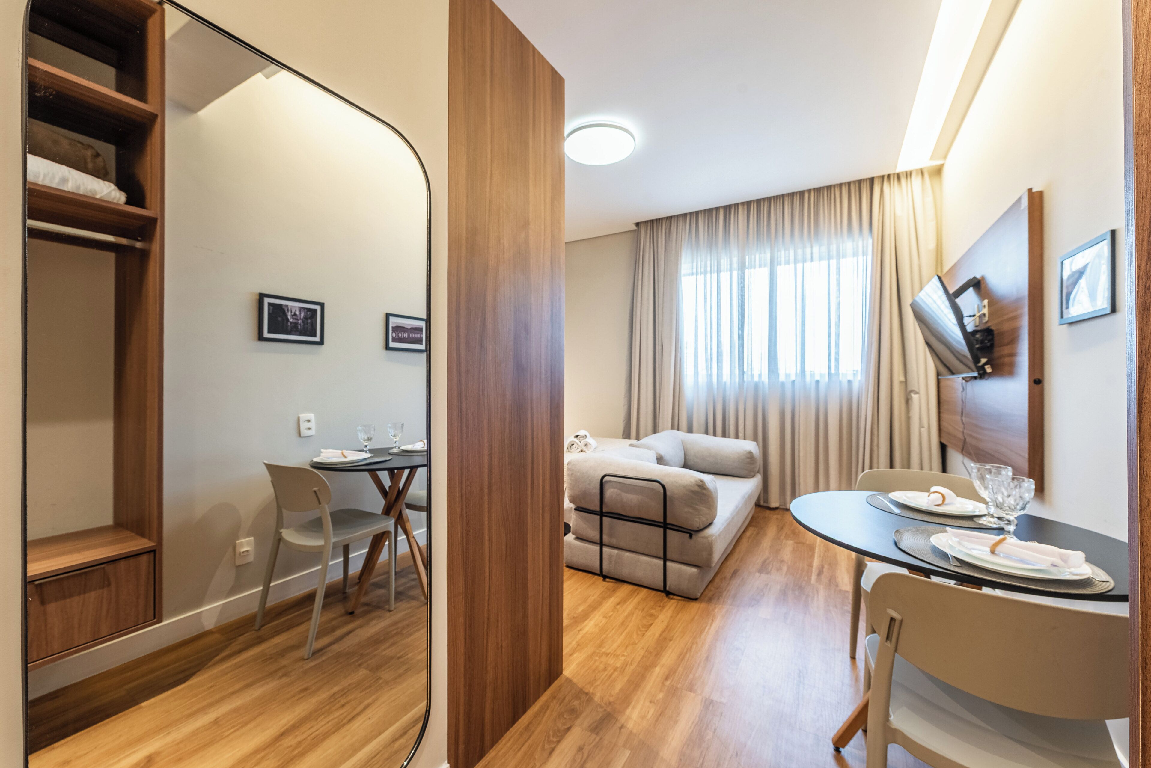 Une chambre d’hôtel moderne avec un plancher en bois, un canapé, une table à manger avec des chaises, un téléviseur fixé au mur et une fenêtre avec des rideaux.