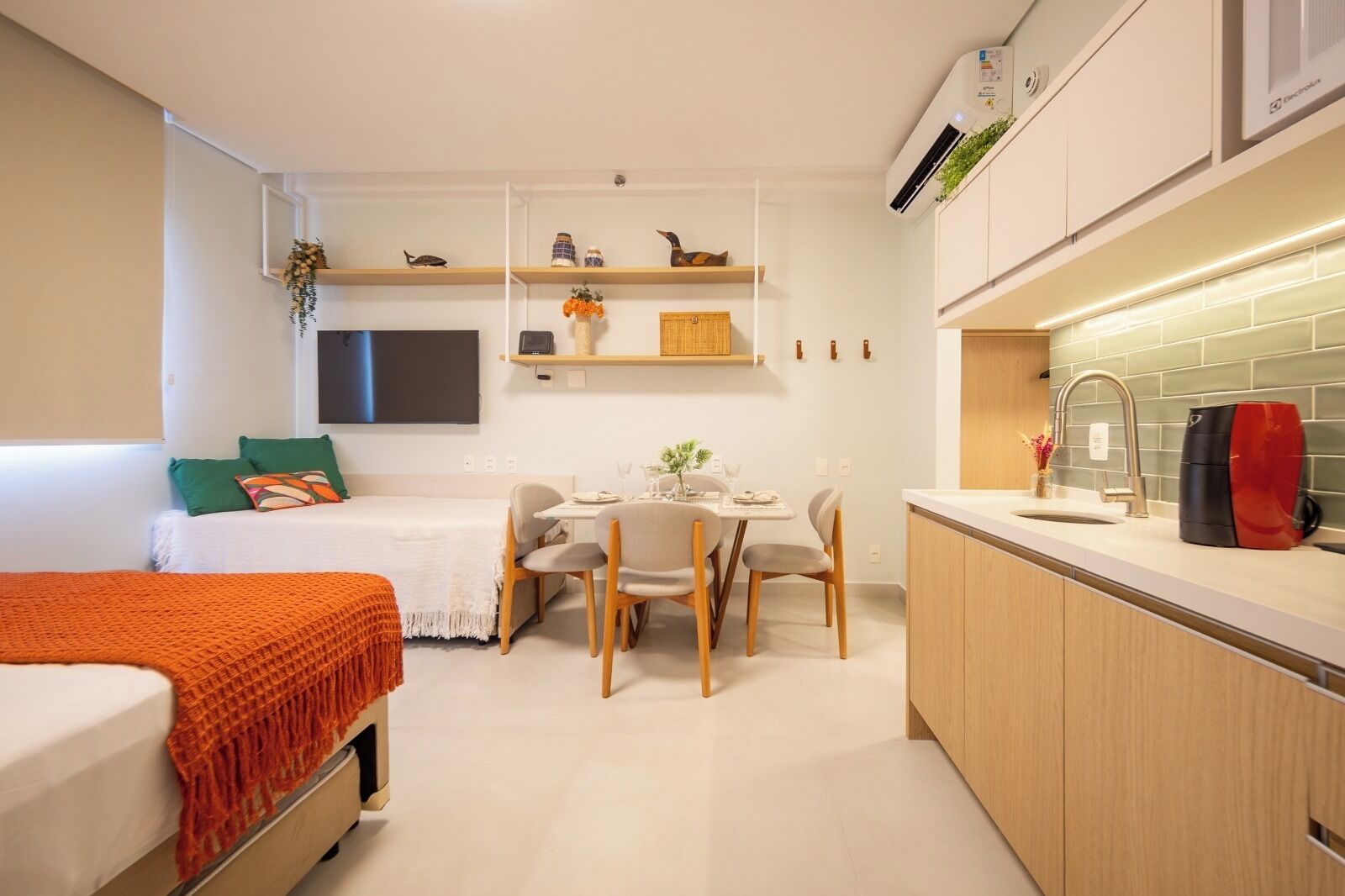 A compact living space with a bed, a dining area, a kitchen with a sink and a kettle, and a mounted air conditioner.
