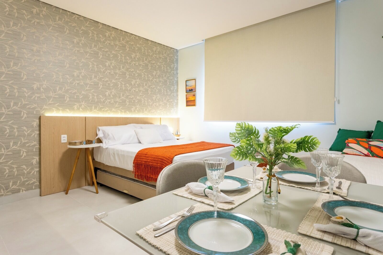 A modern hotel room with a neatly made bed, a dining table set for two, and a small side table with a vase of flowers.