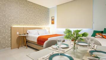 A modern hotel room with a neatly made bed, a dining table set for two, and a small side table with a vase of flowers.