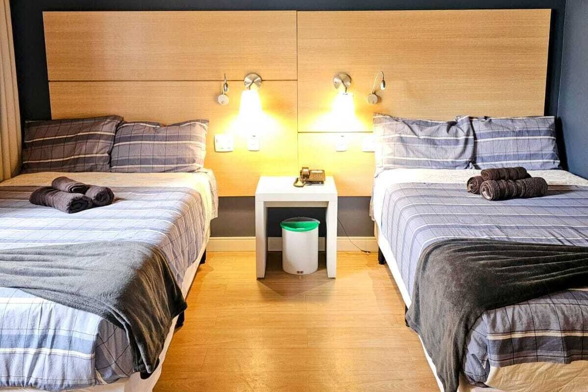 A hotel room with two beds, a nightstand with a lamp, and a trash bin.