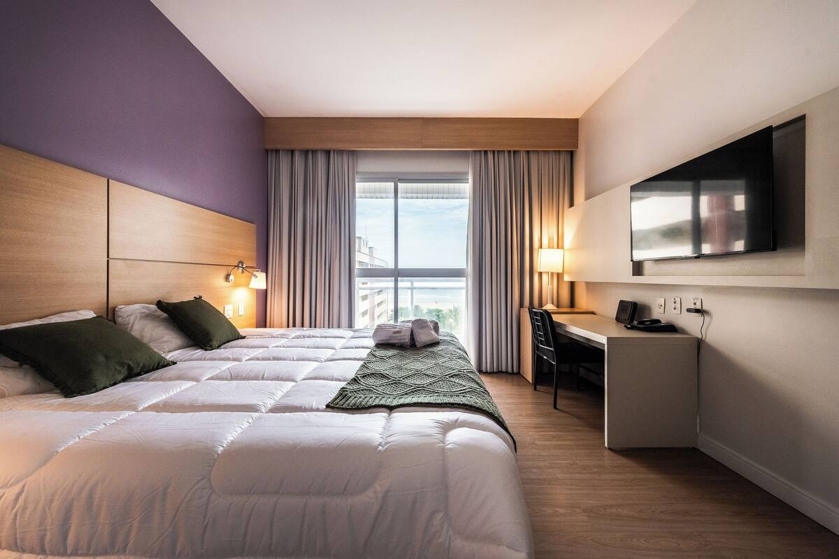 A hotel room with a large bed, a desk, a TV, and a view of the city through the window.