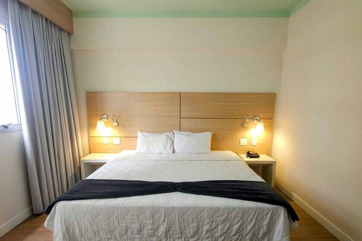 A hotel room with a large bed, wooden headboard, two bedside lamps, and a window with curtains.