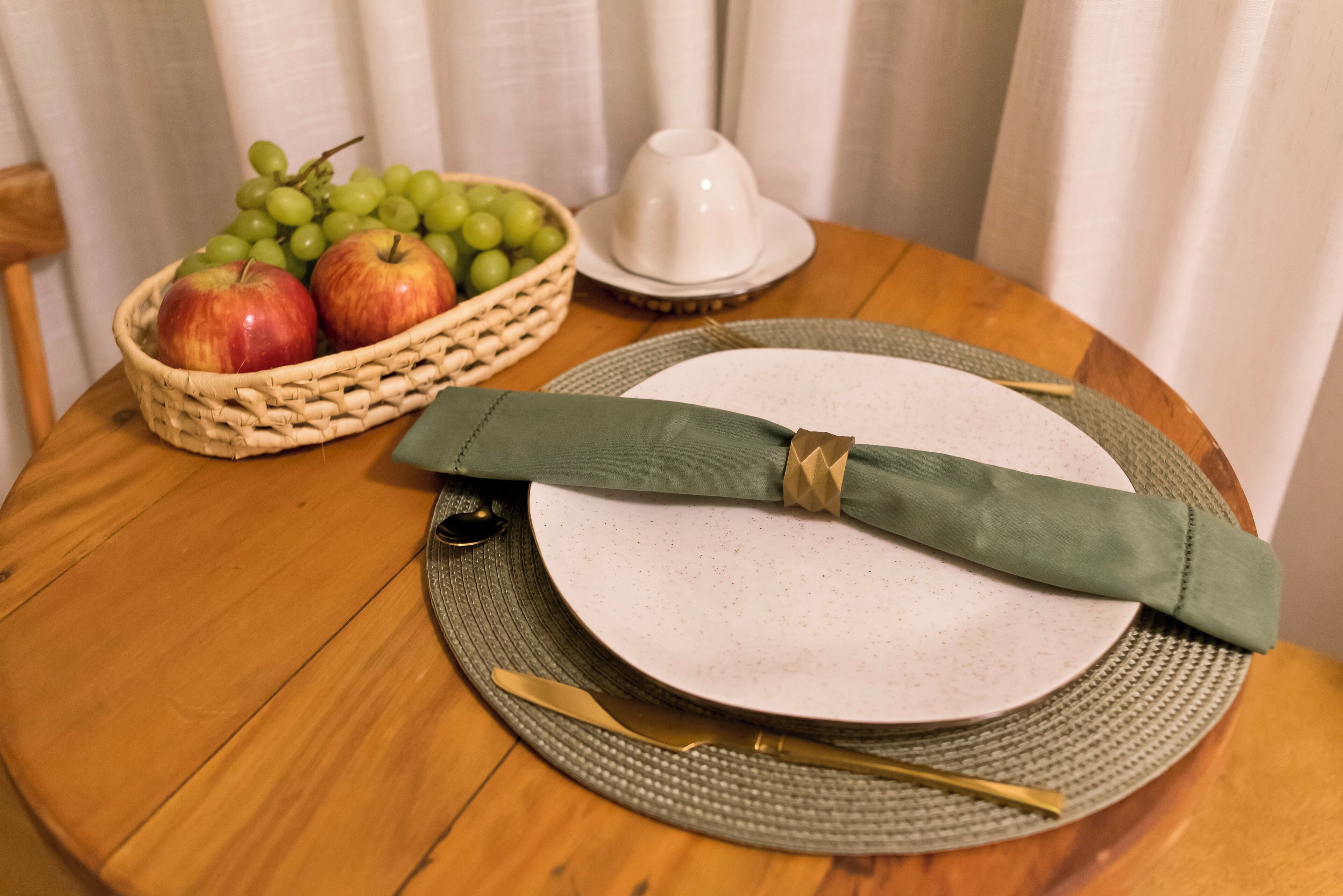 A round wooden table with a white plate, a green napkin, a gold spoon, and a wicker basket containing apples and grapes.