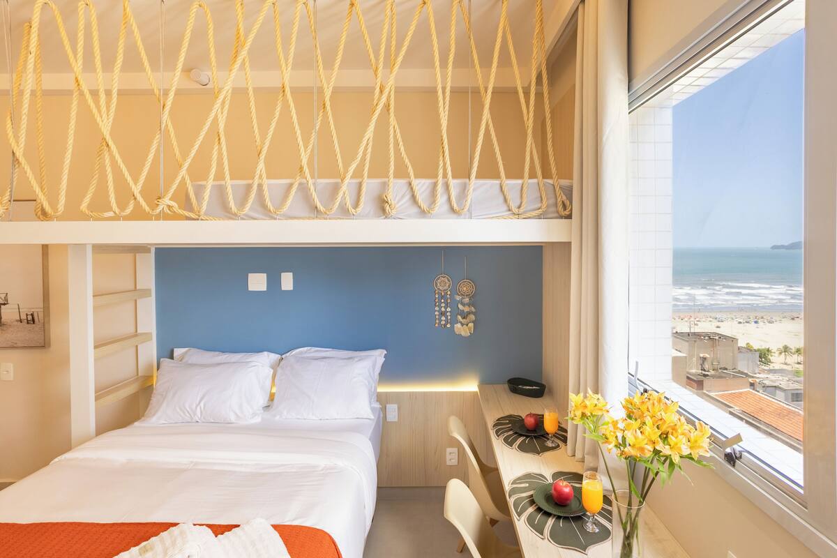 A hotel room with a bed, a desk with a laptop, a vase with flowers, and a view of the beach.