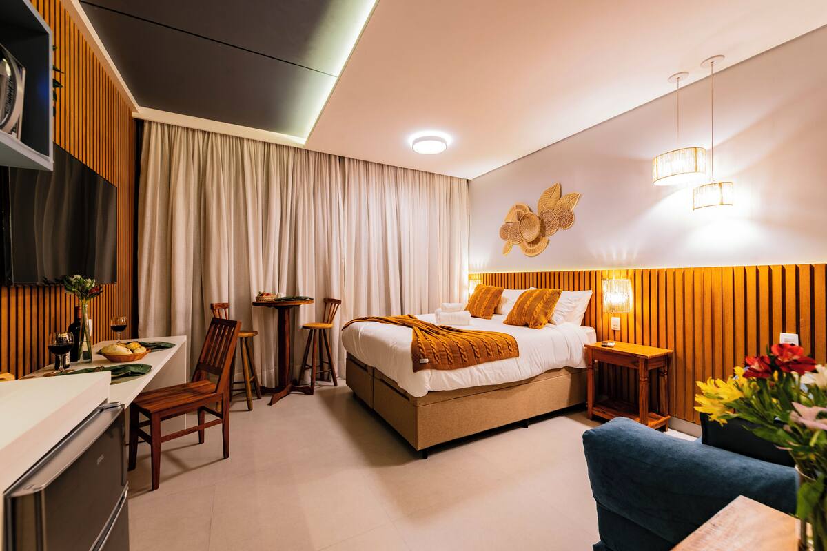 A modern hotel room with a bed, bedside table, chair, and a small dining area with a table and chairs.