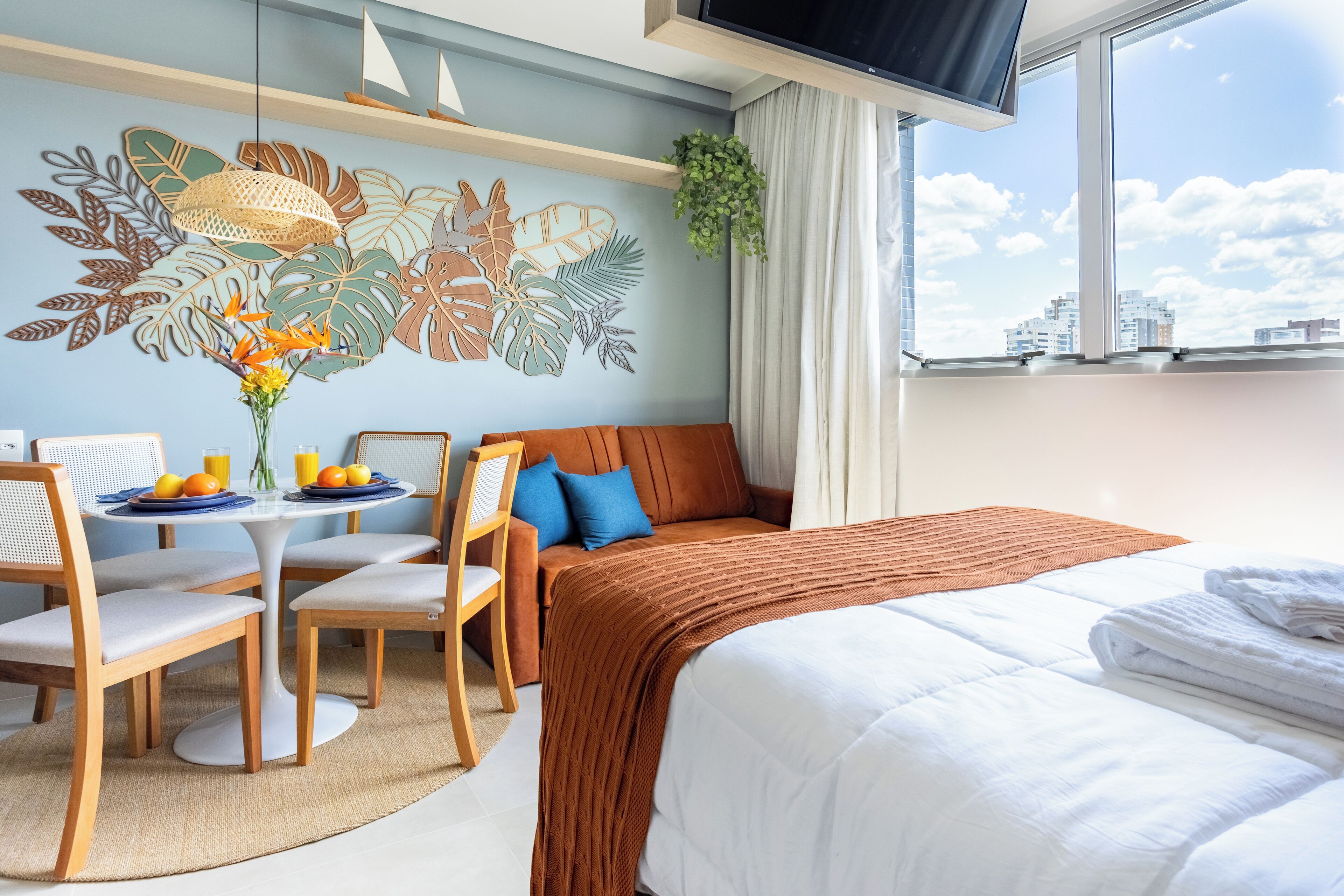 Hotellihuone, jossa on sänky, sohva, ruokapöytä tuoleineen, seinämaalaus ja näkymä rakennuksiin ikkunasta.