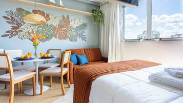 A hotel room with a bed, a sofa, a dining table with chairs, a wall mural, and a view of buildings through the window.