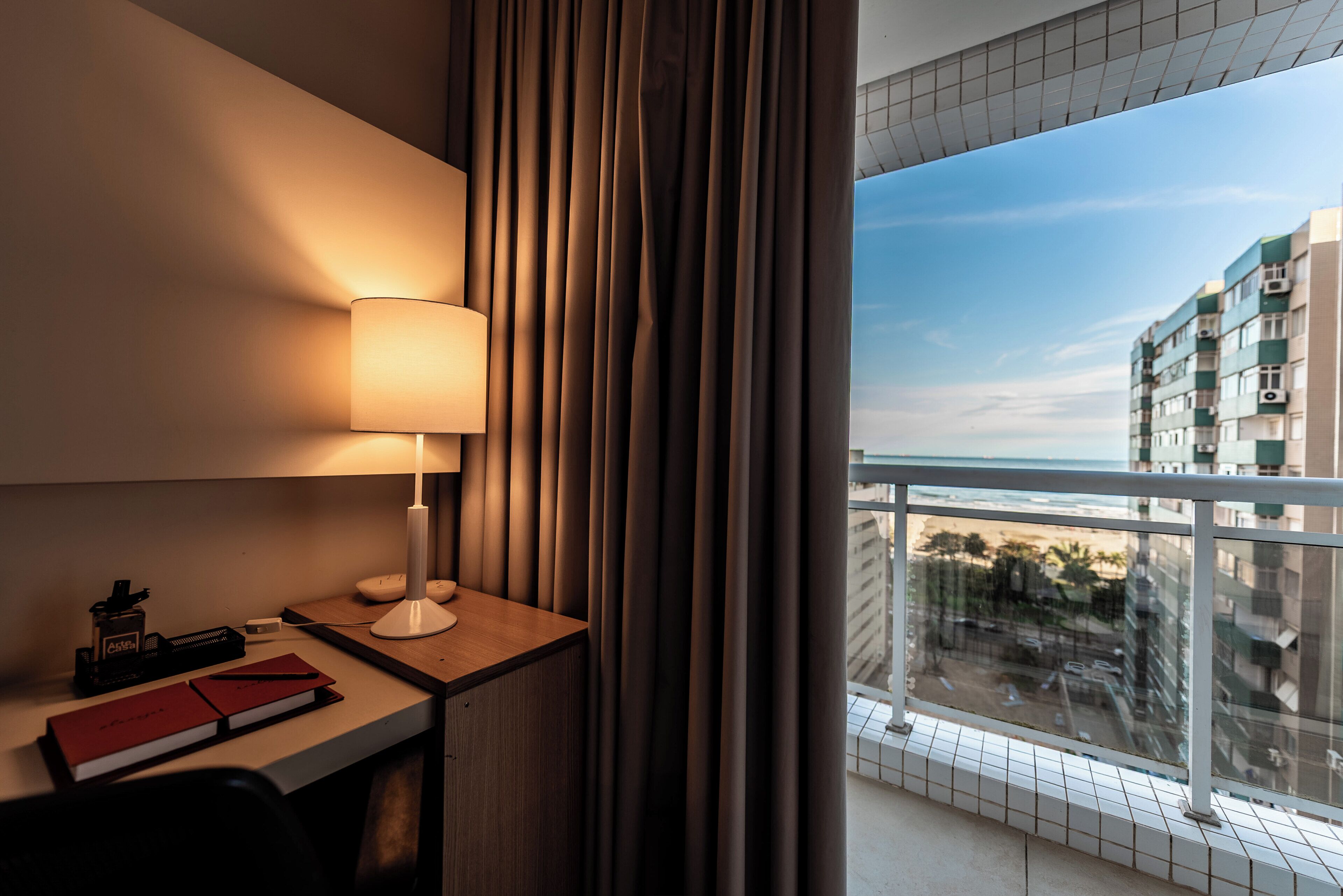 Habitación de hotel con escritorio, lámpara y vistas de la ciudad desde un balcón.