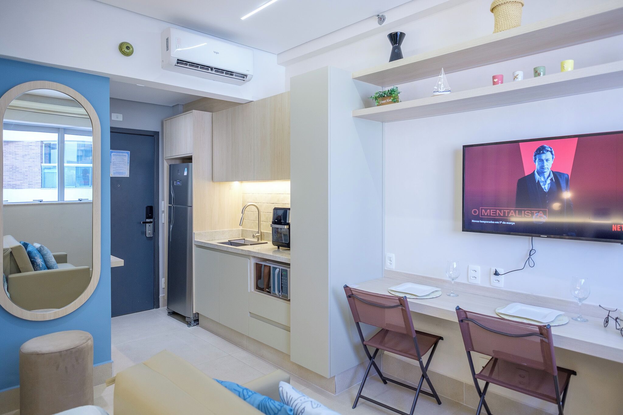 Una cocina moderna con un refrigerador empotrado, un área de comedor con dos sillas y un televisor de pantalla plana montado en la pared mostrando un programa.