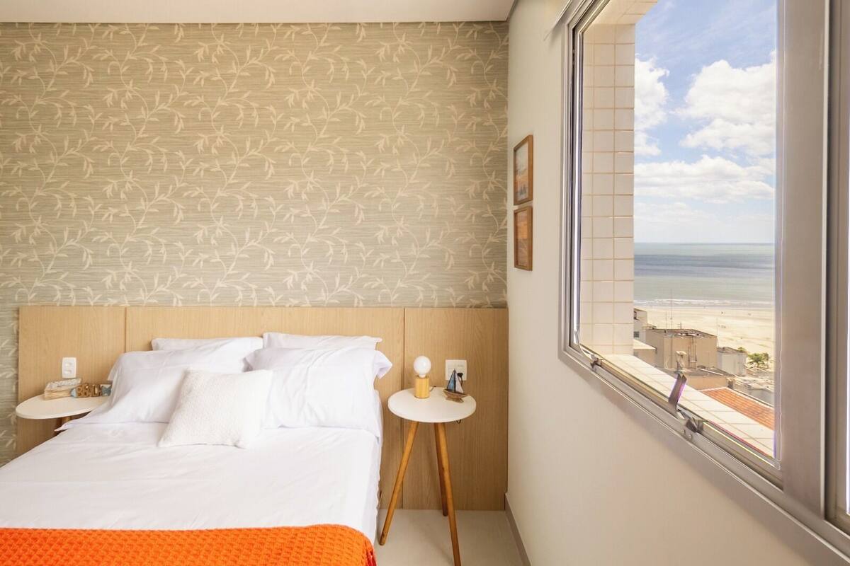 A hotel room with a bed, a small round table with a lamp, a view of the beach, and a patterned wallpaper.