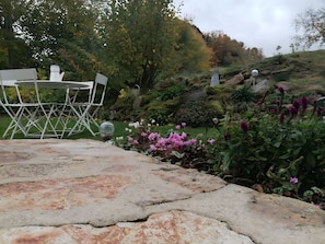 Un patio in pietra con tavolo e sedie in metallo, immerso in un giardino ricco di piante e fiori.