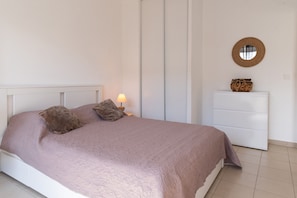 Una cama bien hecha con cabecera blanca, una lámpara de noche y una pequeña cesta de madera sobre un tocador blanco.