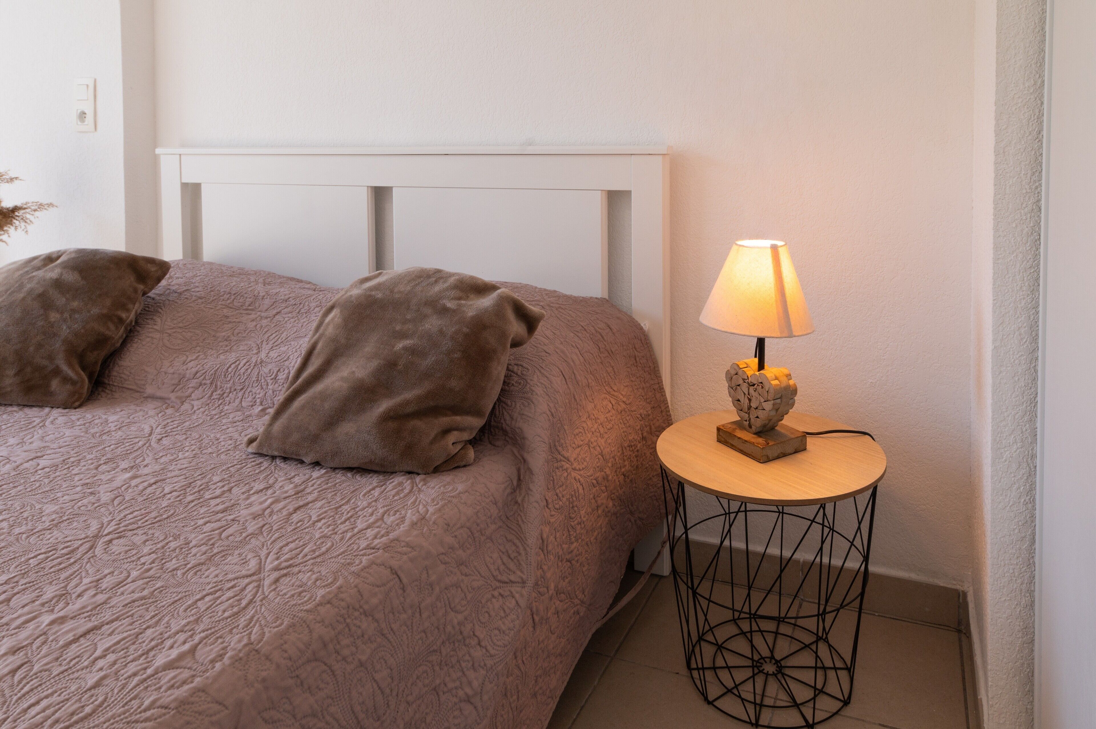 Un lit bien fait, avec une couverture à motifs et deux oreillers, une lampe de chevet posée sur une petite table ronde et une tête de lit blanche.