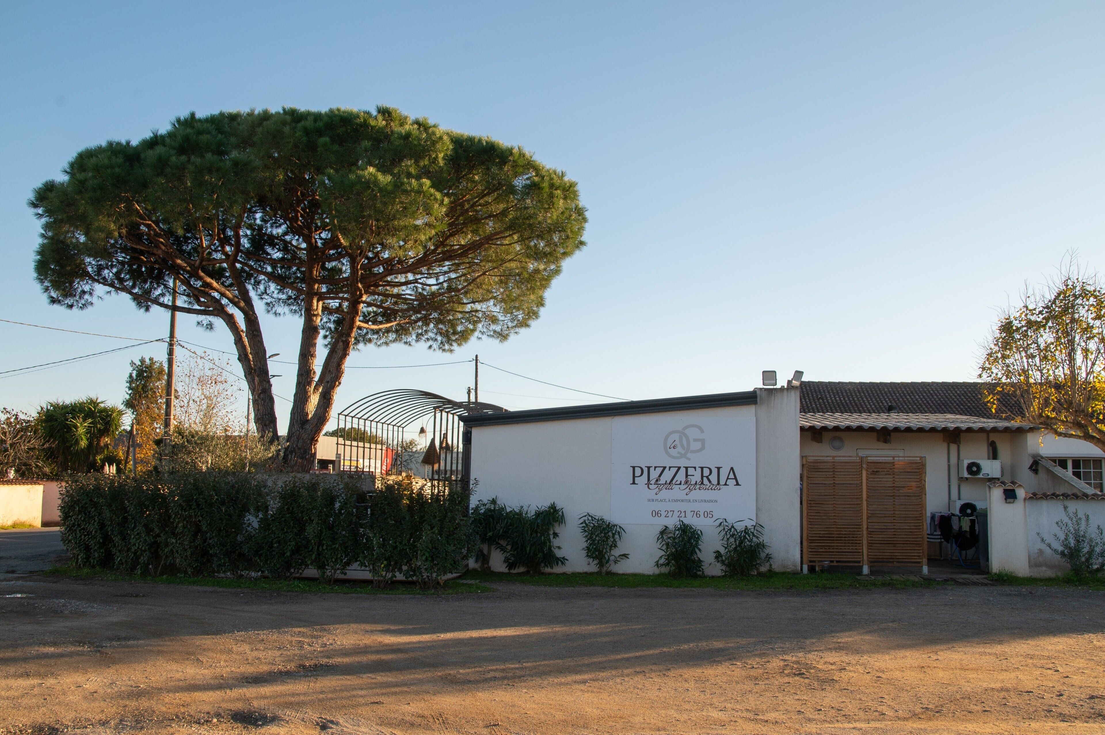 Een pizzeria met een witte gevel, een grote boom en een metalen hek.