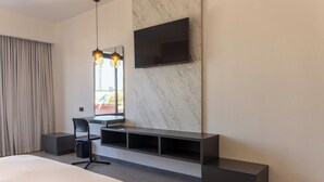 A modern hotel room with a large bed, a desk with a chair, a TV mounted on a marble wall, and a window with curtains. - MARUVA Hotel Ejecutivo (Tecomán)