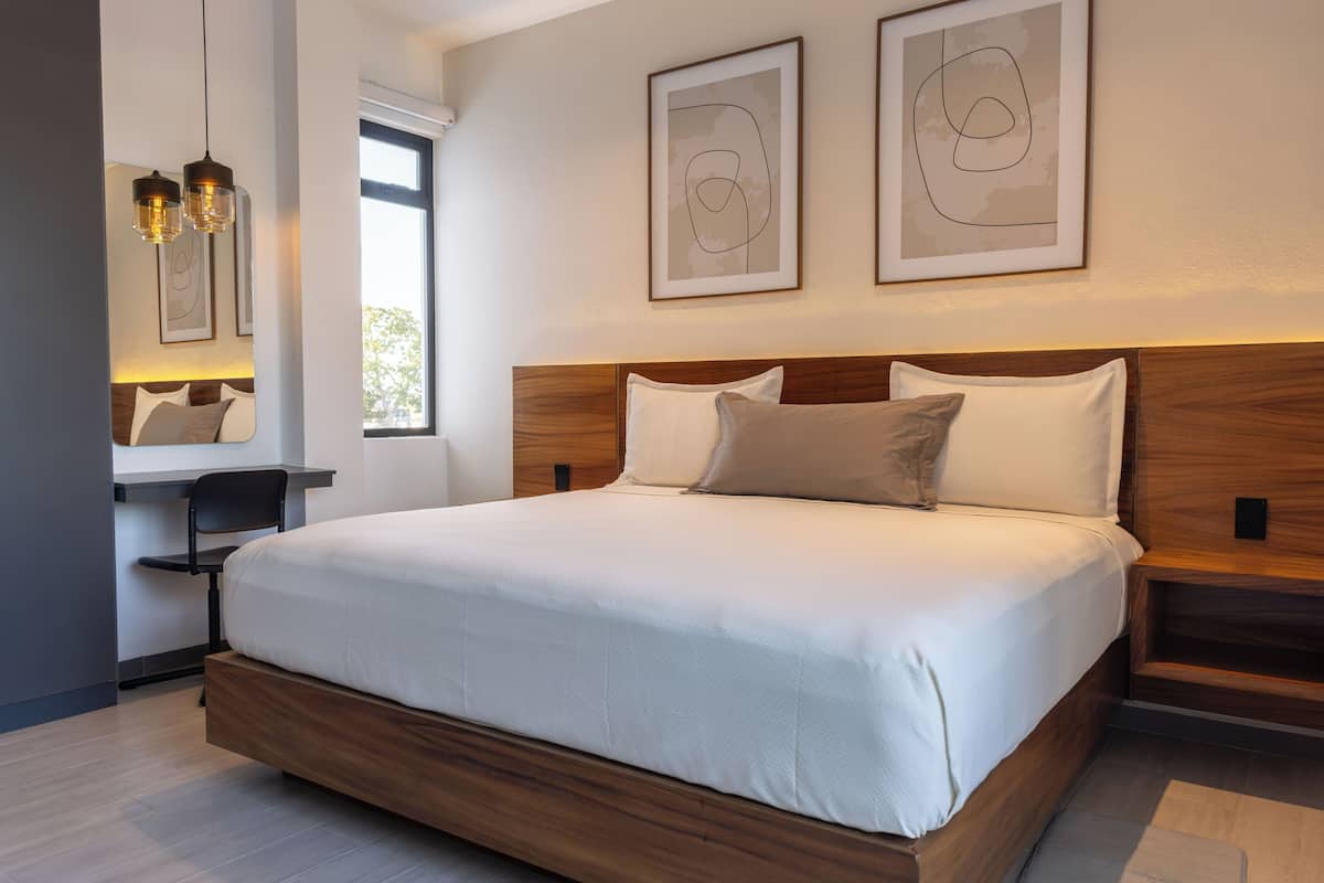 Ein modernes Hotelzimmer mit einem großen Bett, einem Schreibtisch und einem Stuhl. An den Wänden befinden sich abstrakte Gemälde und ein Fenster lässt natürliches Licht herein.