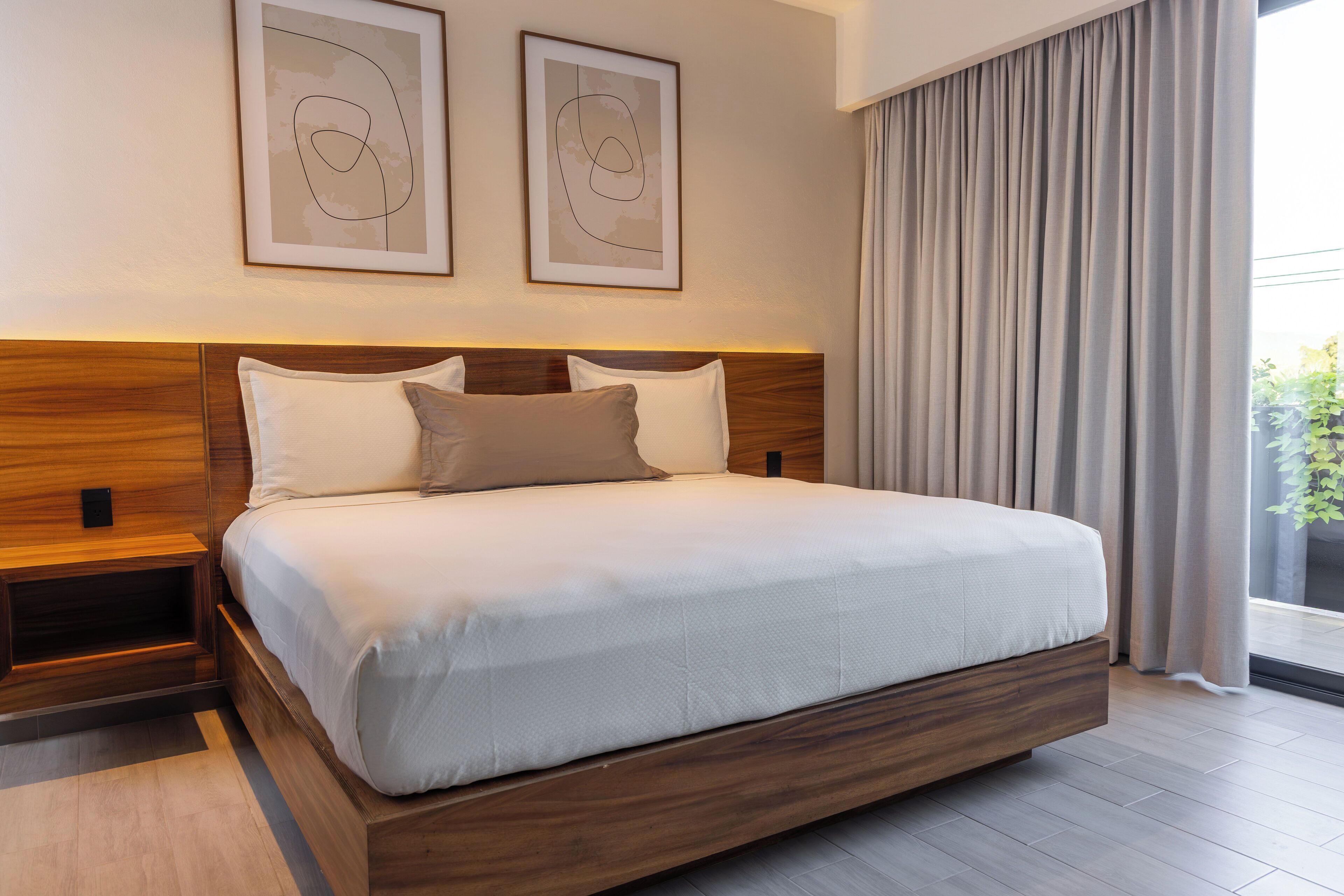 Un dormitorio moderno con cabecera de madera, una cama grande con ropa de cama blanca, dos obras de arte abstracto enmarcadas y vistas al exterior con vegetación a través de una ventana con cortinas.