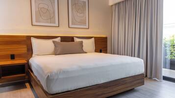 Un dormitorio moderno con cabecera de madera, una cama grande con ropa de cama blanca, dos obras de arte abstracto enmarcadas y vistas al exterior con vegetación a través de una ventana con cortinas.