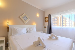 Une chambre d’hôtel avec un grand lit, du linge de lit blanc, un tableau encadré au mur, une table de chevet et une fenêtre avec des rideaux.