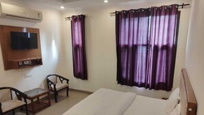 A hotel room with a bed, a TV mounted on the wall, two chairs, a small table, and purple curtains. - Hotel Atlantiis Suites by Signature Stays (New Delhi)