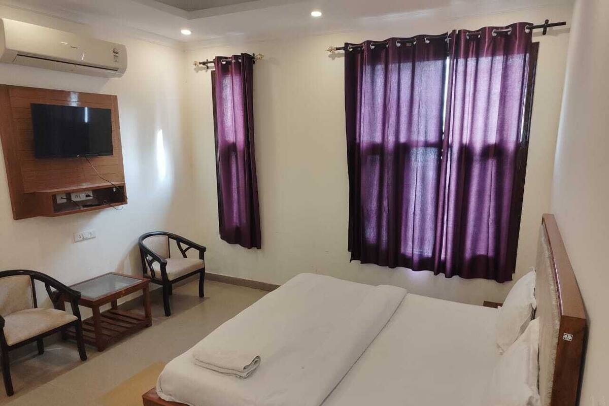 Habitación de hotel con cama, televisor montado en la pared, dos sillones, una mesita y cortinas moradas.