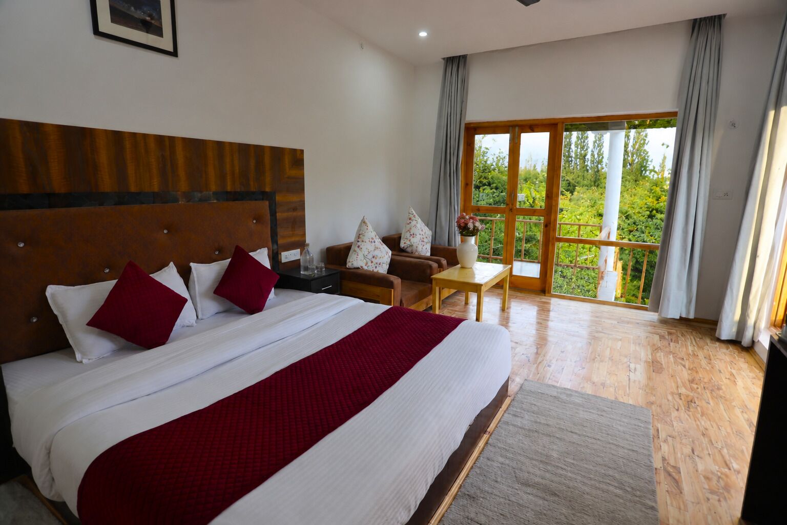 A hotel room with a large bed, a wooden headboard, a sofa, a small table, and a balcony with a view of greenery.