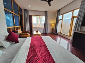 A hotel room with a large bed, a wooden chair, a small table with a lamp, and large windows offering a view of the mountains. - Indus Serenity Retreat, Ladakh (Leh)