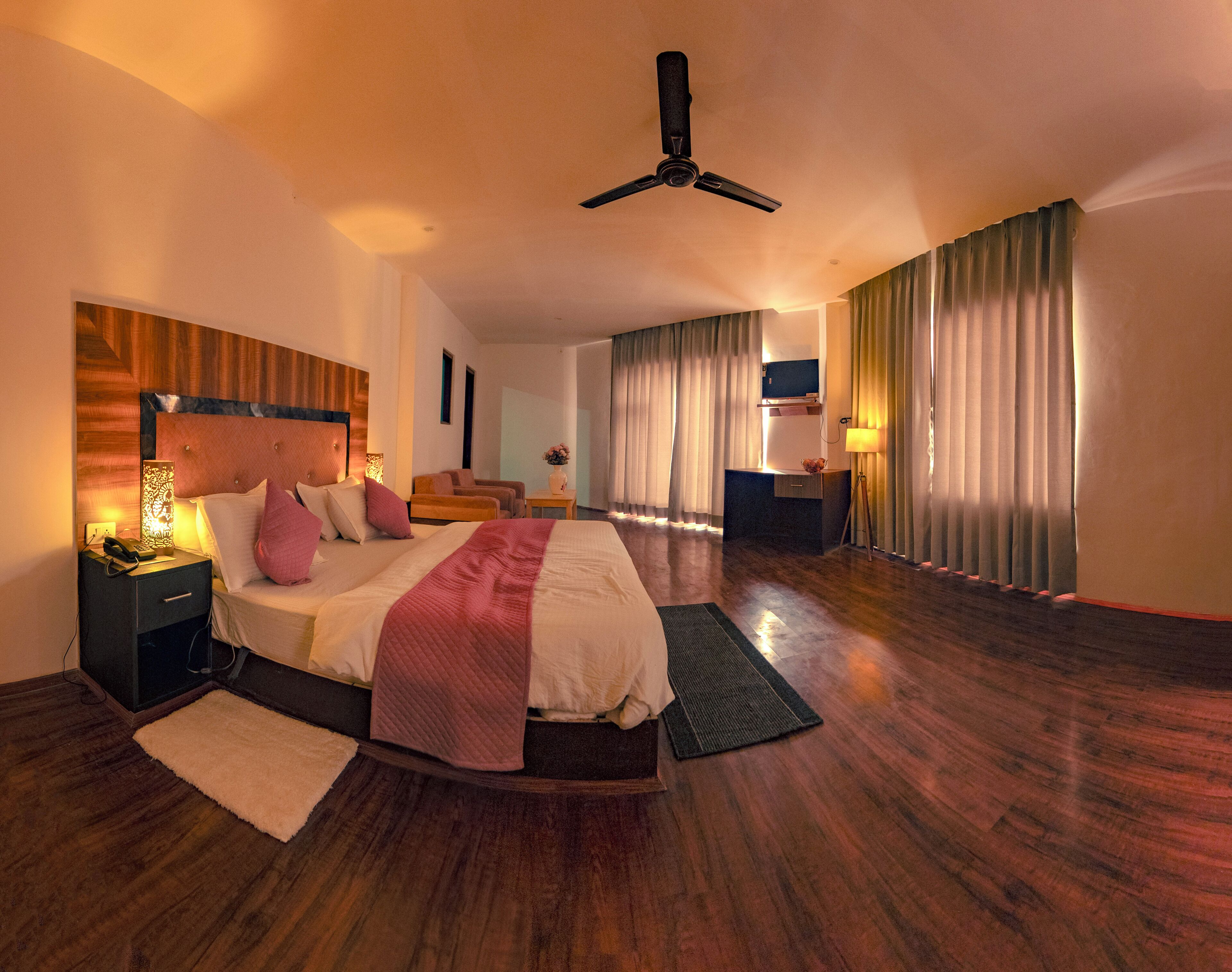 A spacious hotel room with a large bed, a desk, a chair, a standing lamp, and a ceiling fan.