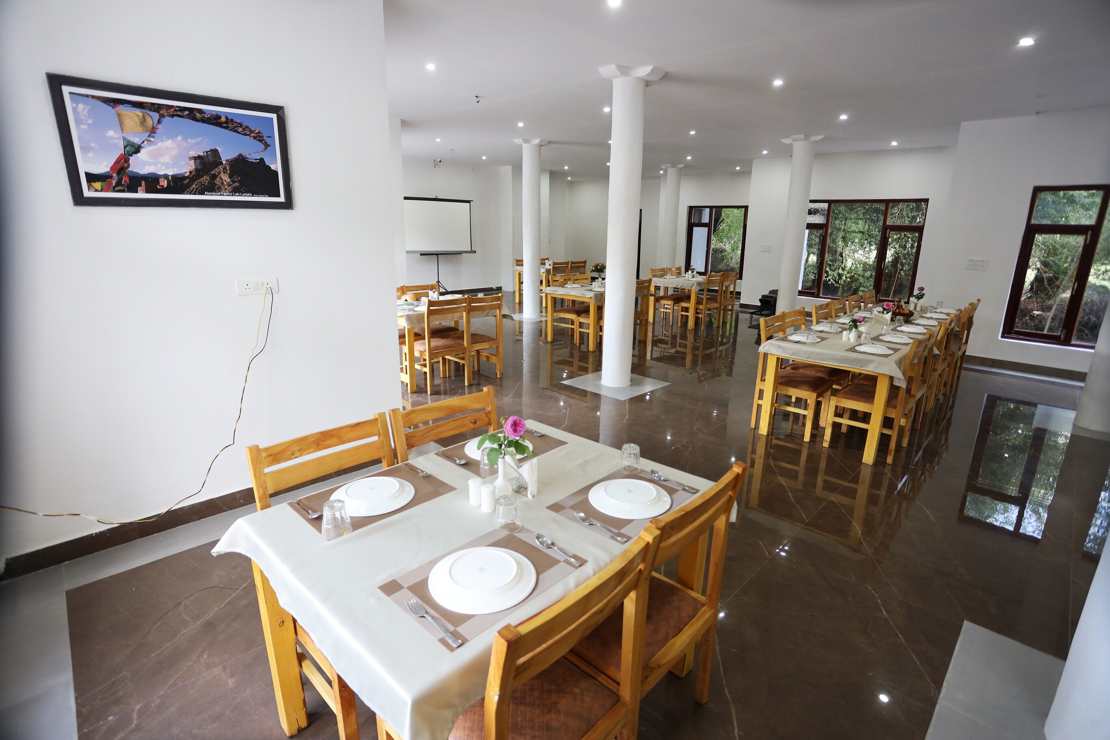 Un comedor con varias mesas dispuestas para comensales, una pantalla de proyector en la pared y un cuadro colgado sobre las mesas.