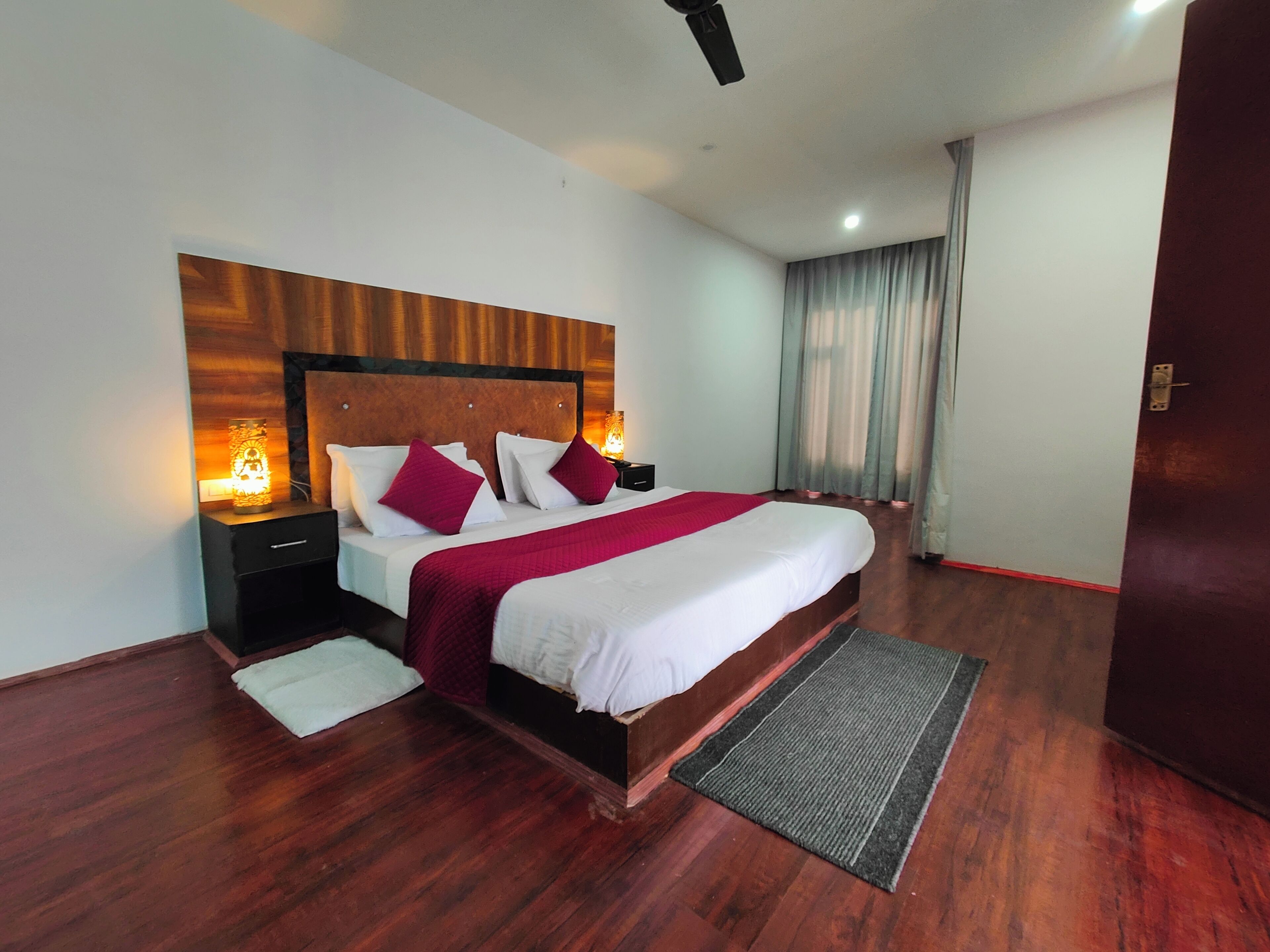 A modern hotel room with a wooden headboard, a large bed with white bedding and red accents, a bedside table with a lamp, and a view of the outside through a window with curtains.
