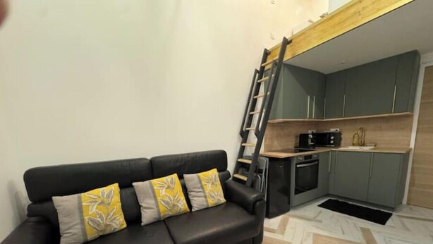 A modern living room with a black sofa and patterned cushions, a herringbone-patterned floor, and a kitchen area with grey cabinets and a wooden countertop. - Self-contained studio near Paris (Vitry-sur-Seine)