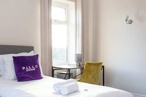 Ein Hotelzimmer mit einem Bett, einem violetten Kissen, einem gelben Sessel, einem Schreibtisch mit Lampe und einem Fenster mit Vorhängen.