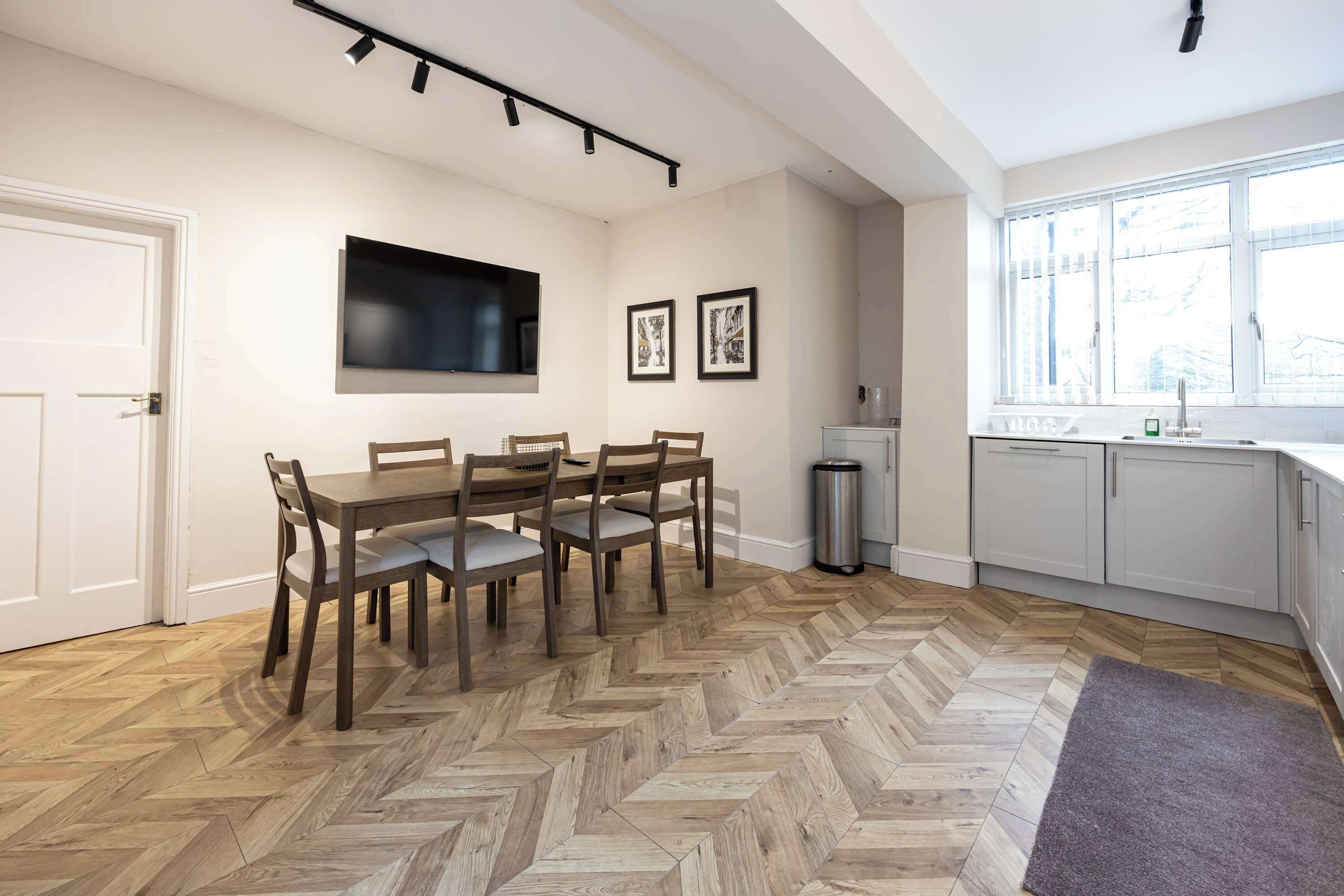 A modern kitchen with a herringbone wood floor, white cabinets, a large window, and a dining area with a wooden table and chairs.