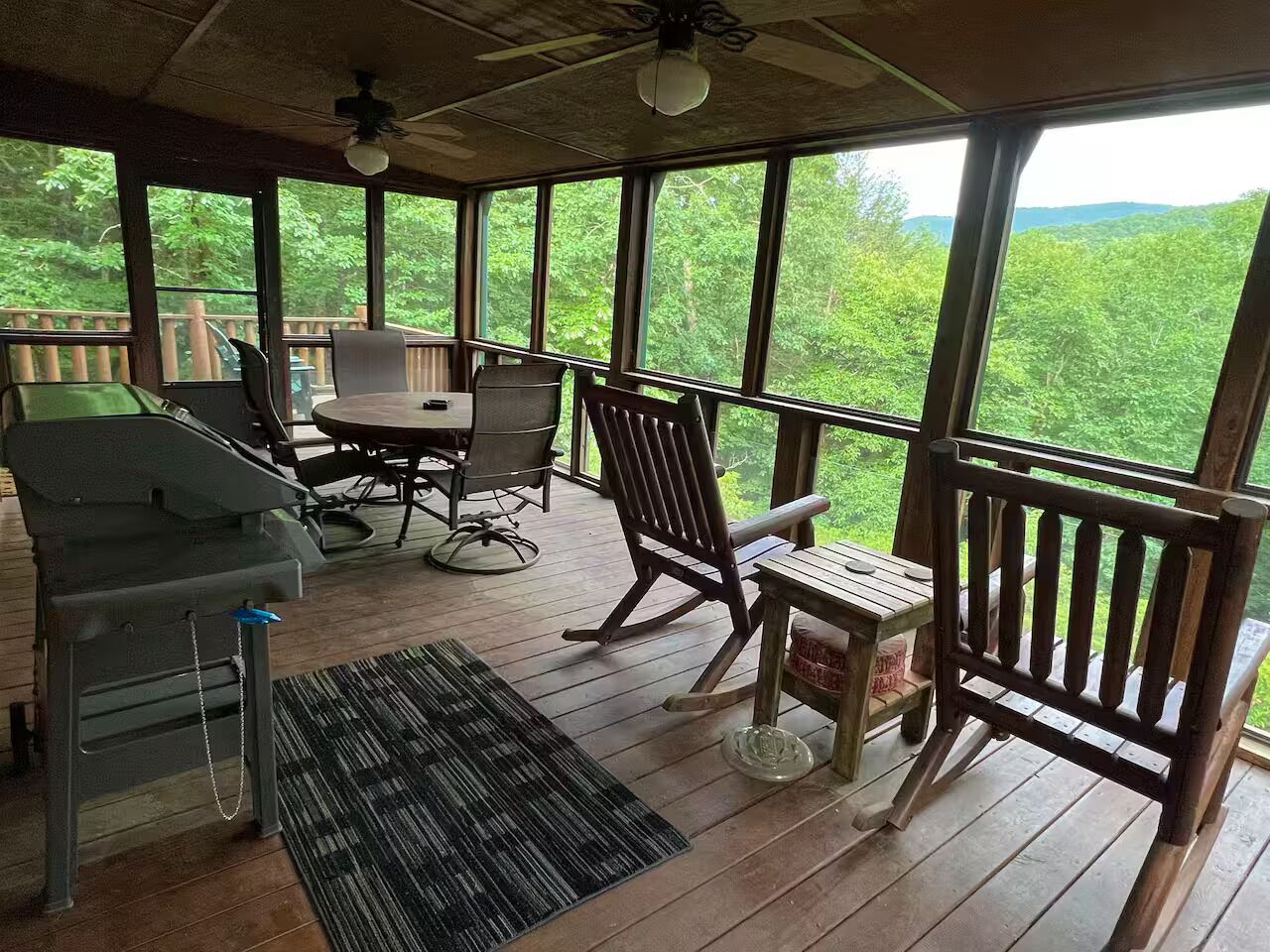 A covered outdoor area with a grill, dining table, chairs, and a view of greenery.