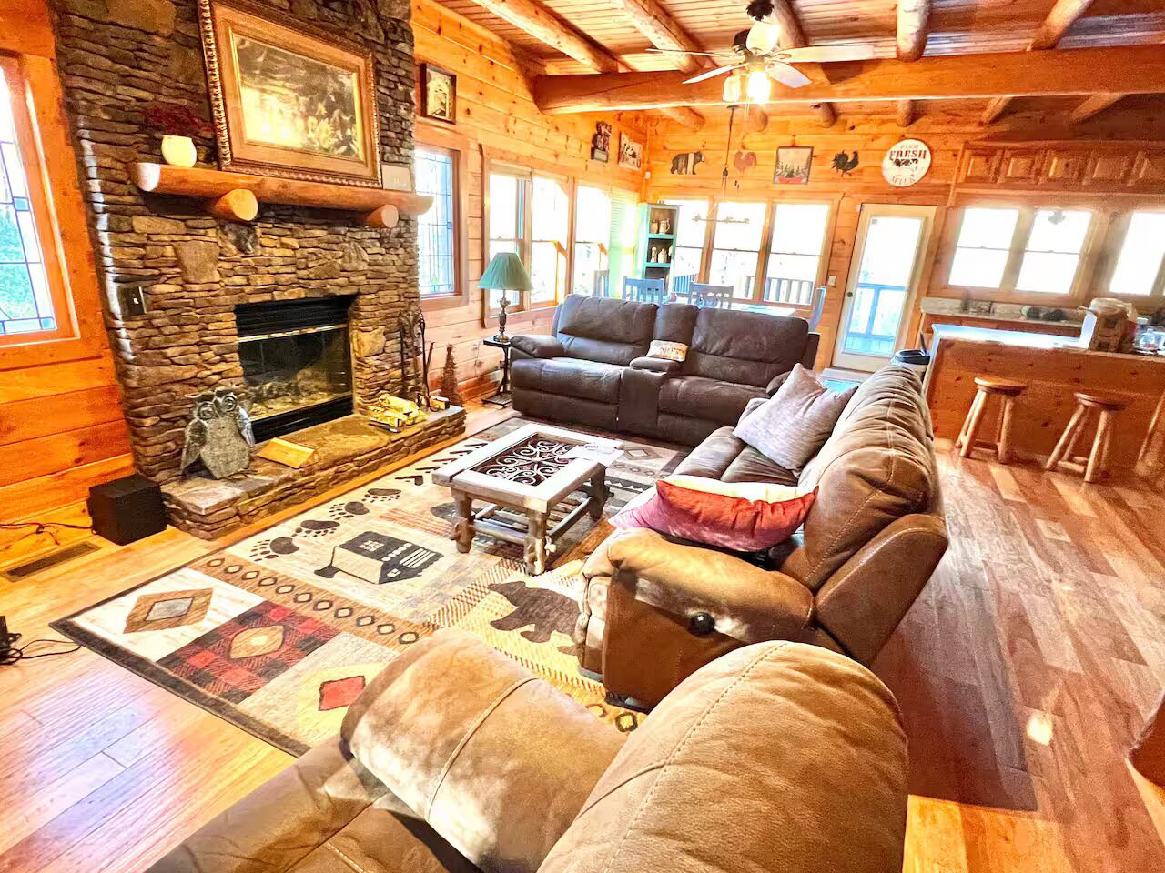 A cozy log cabin interior with a stone fireplace, leather sofas, and a patterned rug.