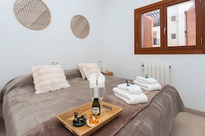 A hotel room with a bed, two pillows, a tray with a bottle and chocolates, and a window with blinds.