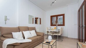 A living room with a brown sofa, white cushions, a small table with a lamp, and a dining area with a table and chairs.