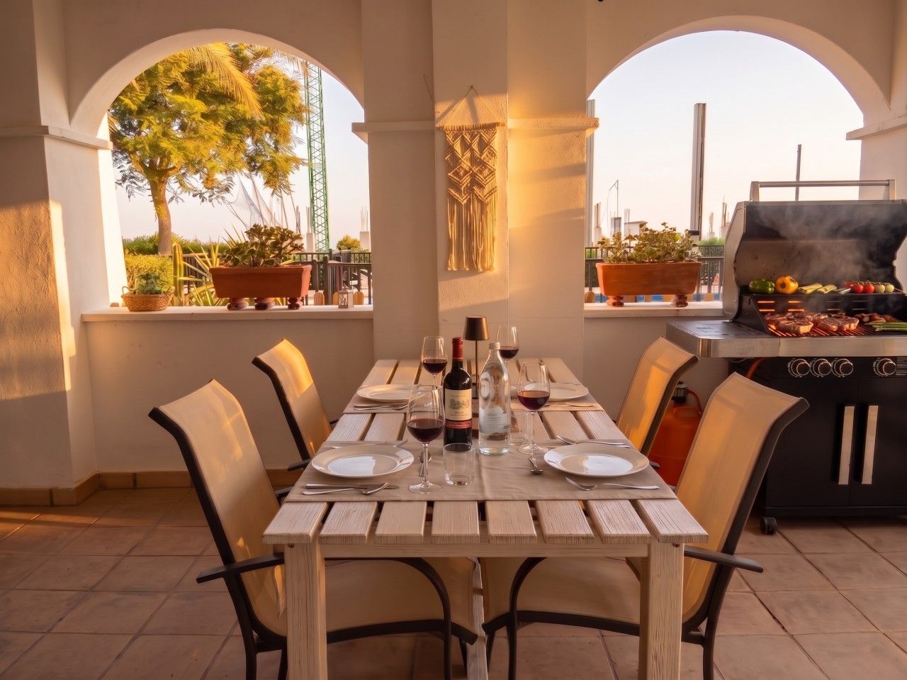 An outdoor dining area with a table set for four, surrounded by chairs, and a grill in the background.