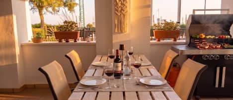 An outdoor dining area with a table set for four, surrounded by chairs, and a grill in the background.