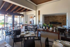 A spacious dining area with multiple tables and chairs, a fireplace, and large windows allowing natural light.