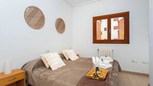 A bedroom with a bed, pillows, a bedside table, a tray with toiletries, and a window with blinds.