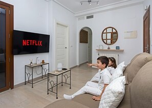 Een woonkamer met een flatscreen-tv waarop 'NETFLIX' staat, een bank, een salontafel en een deur naar een andere kamer.