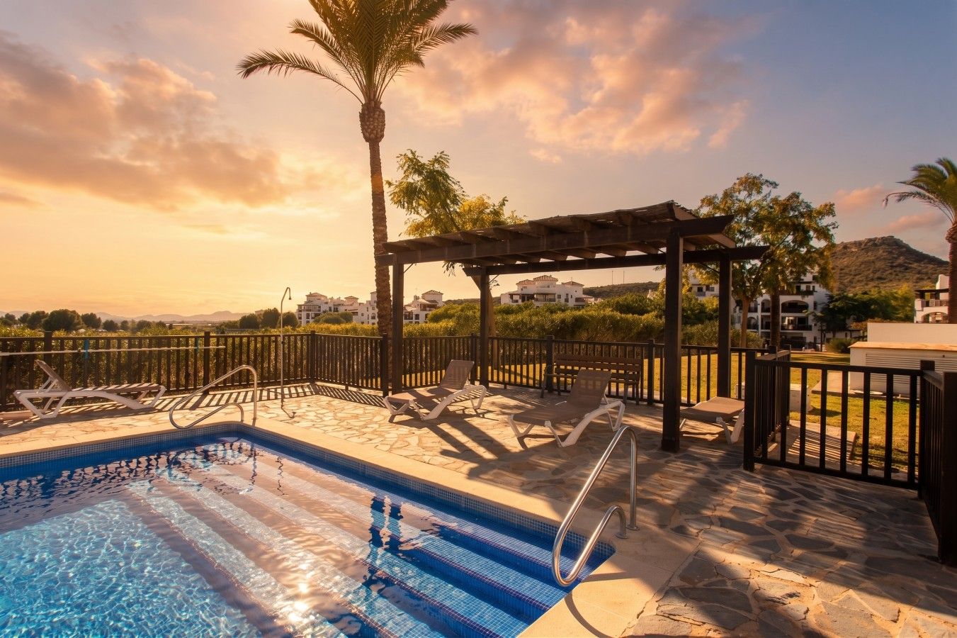 A swimming pool with lounge chairs and a pergola, surrounded by a fence and palm trees.