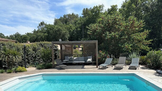 Een zwembad met een houten terras, ligbedden en een houten pergola met kussens.