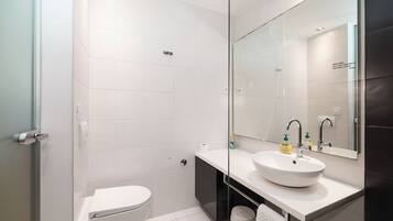 Un baño moderno con inodoro blanco, cabina de ducha con vidrio, lavamanos blanco y un espejo grande.