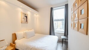 Um quarto com cama, mesa de cabeceira, luminária, janela com cortinas e uma obra de arte na parede.