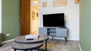 Uma sala de estar com televisão de tela plana, uma mesa redonda com um livro e um controle remoto, um armário cinza e um tapete estampado.